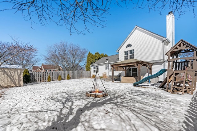 snow covered back of property featuring a playground, a fenced backyard, a fire pit, a gazebo, and a chimney