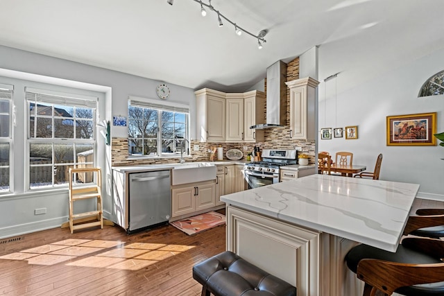 kitchen with lofted ceiling, light stone counters, stainless steel appliances, wall chimney range hood, and a sink