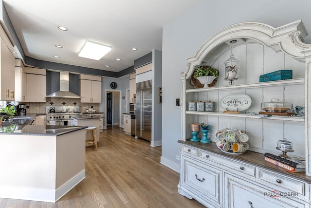 kitchen featuring high end range, dark countertops, wall chimney exhaust hood, light wood-type flooring, and backsplash