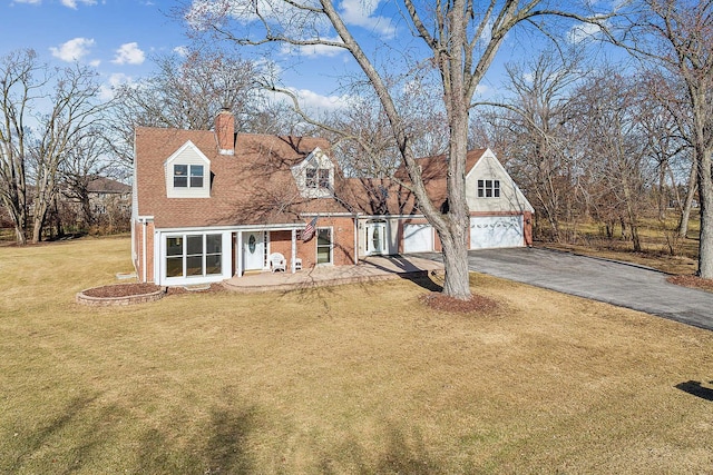 new england style home featuring driveway, brick siding, a chimney, and a front yard