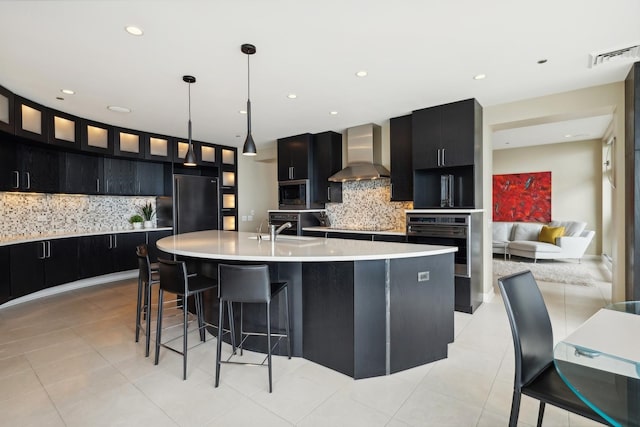 kitchen featuring pendant lighting, tasteful backsplash, black appliances, an island with sink, and wall chimney exhaust hood