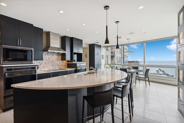 kitchen with black appliances, decorative light fixtures, an island with sink, wall chimney range hood, and backsplash