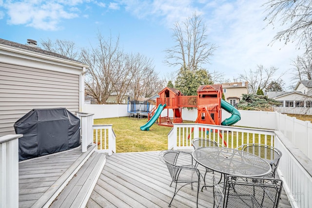 wooden deck featuring a trampoline, outdoor dining area, a playground, a yard, and a fenced backyard