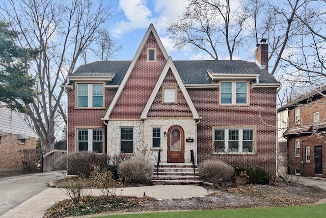 english style home with brick siding, a chimney, a shingled roof, entry steps, and stone siding