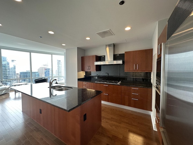 kitchen featuring a city view, appliances with stainless steel finishes, a kitchen island with sink, a sink, and wall chimney exhaust hood