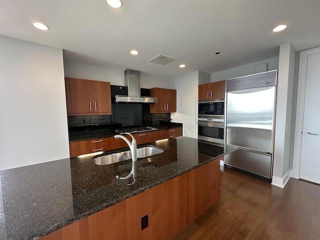 kitchen featuring dark wood-type flooring, a sink, built in appliances, dark stone counters, and wall chimney exhaust hood