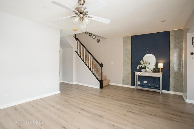 unfurnished living room with an accent wall, ceiling fan, wood finished floors, baseboards, and stairs