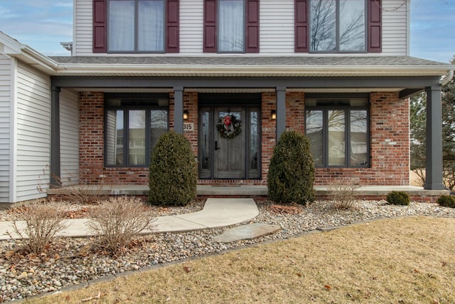 view of exterior entry featuring brick siding, a porch, and roof with shingles