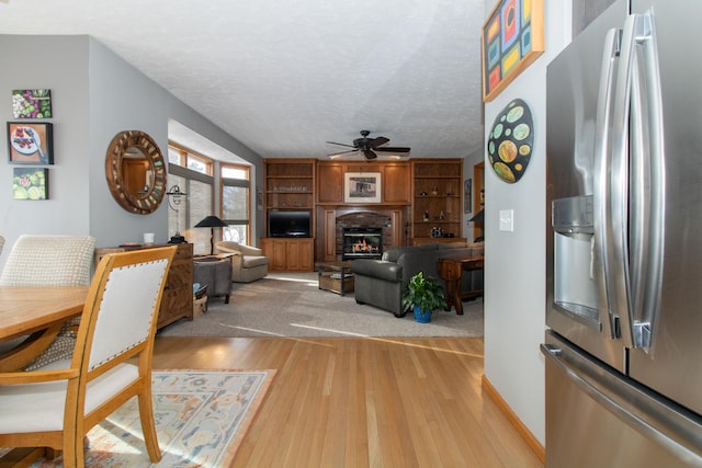 living area with a brick fireplace, a ceiling fan, light wood finished floors, and a textured ceiling