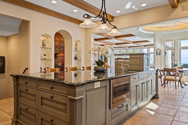 kitchen featuring stainless steel microwave, hanging light fixtures, a kitchen island with sink, a sink, and dark stone counters