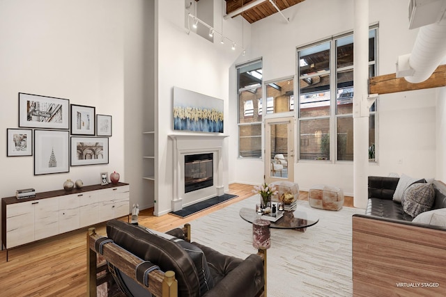 living room featuring a high ceiling, light wood-style floors, beam ceiling, a glass covered fireplace, and rail lighting