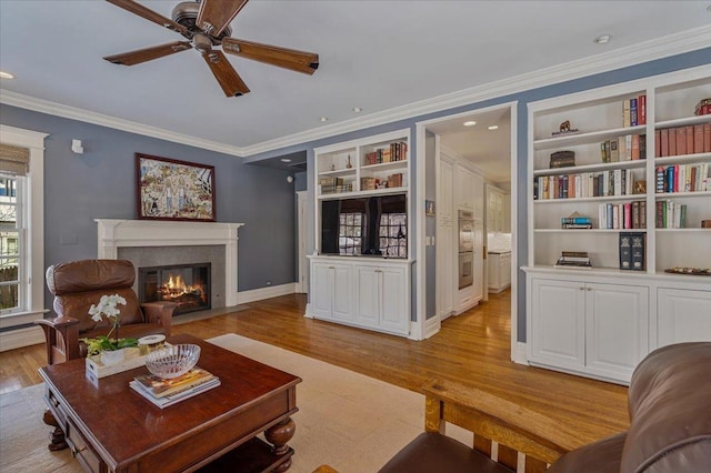 living room with ceiling fan, a fireplace with flush hearth, built in features, ornamental molding, and light wood-type flooring