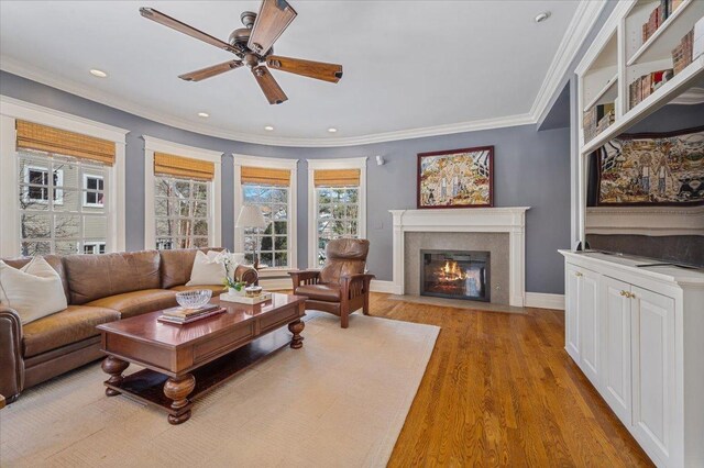 living room featuring baseboards, a ceiling fan, a fireplace with flush hearth, light wood-style flooring, and ornamental molding