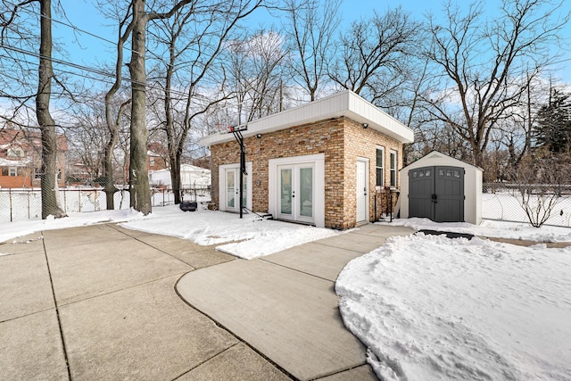 view of snow covered exterior featuring brick siding, an outdoor structure, fence, french doors, and a shed