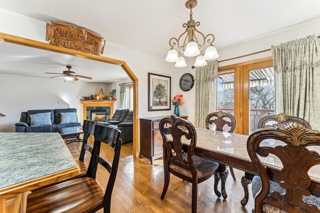 dining room with arched walkways, a fireplace, light wood-style flooring, wainscoting, and ceiling fan with notable chandelier