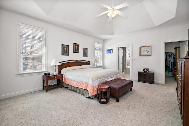 bedroom with connected bathroom, light colored carpet, a walk in closet, ceiling fan, and a raised ceiling