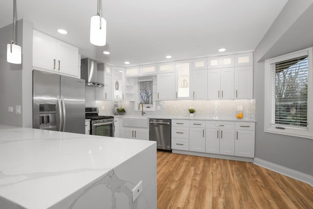 kitchen with appliances with stainless steel finishes, pendant lighting, white cabinets, light stone counters, and wall chimney range hood