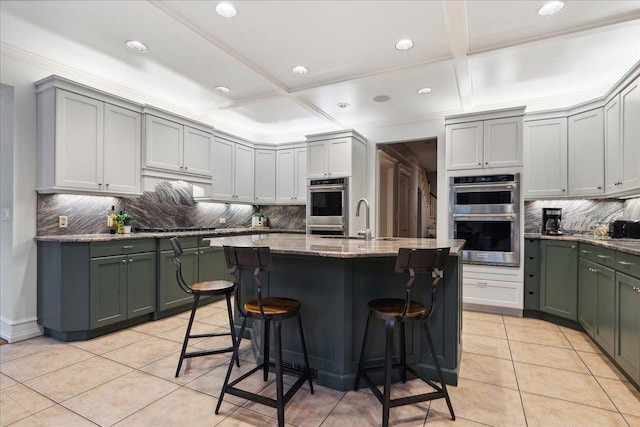 kitchen featuring a center island with sink, double oven, stone counters, light tile patterned flooring, and a breakfast bar