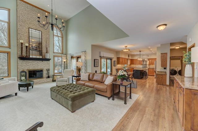 living room featuring a towering ceiling, washing machine and dryer, light wood-type flooring, an inviting chandelier, and a brick fireplace