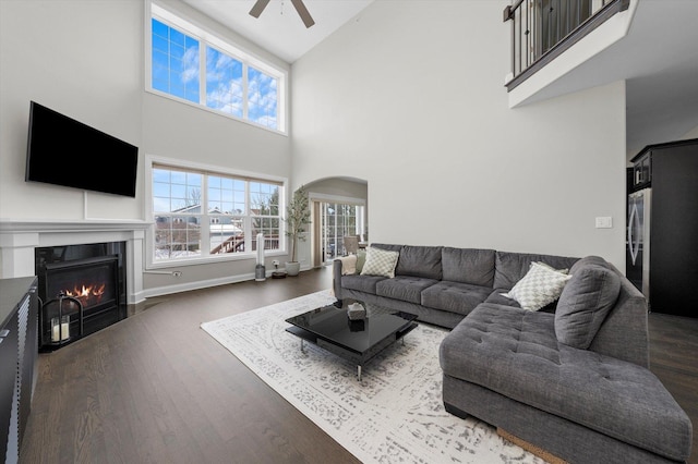 living area with arched walkways, a fireplace with flush hearth, a ceiling fan, baseboards, and dark wood finished floors