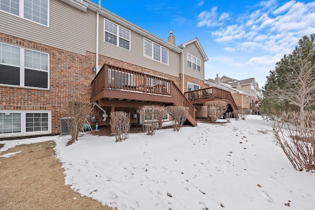 snow covered back of property featuring brick siding, a chimney, and a wooden deck