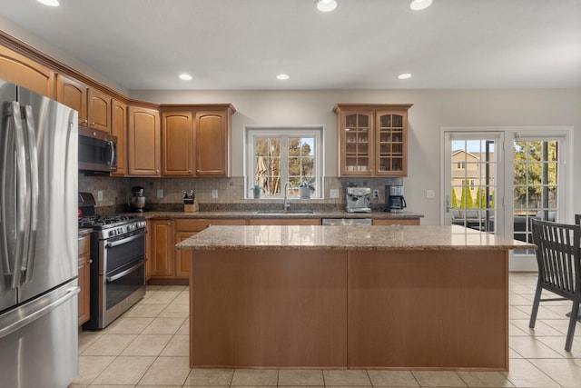 kitchen with light stone counters, stainless steel appliances, a kitchen island, a sink, and glass insert cabinets