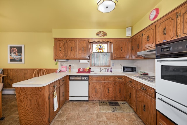 kitchen featuring white appliances, brown cabinetry, a peninsula, under cabinet range hood, and a sink