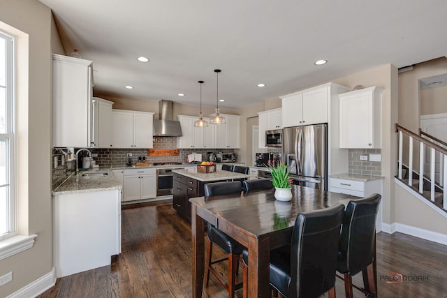 kitchen with decorative light fixtures, stainless steel appliances, a kitchen island, a sink, and wall chimney exhaust hood