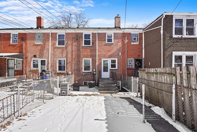 snow covered back of property featuring a fenced front yard, a chimney, cooling unit, and brick siding