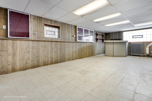 basement featuring radiator heating unit, wooden walls, a drop ceiling, and tile patterned floors