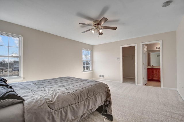 bedroom with connected bathroom, a walk in closet, baseboards, light colored carpet, and a ceiling fan