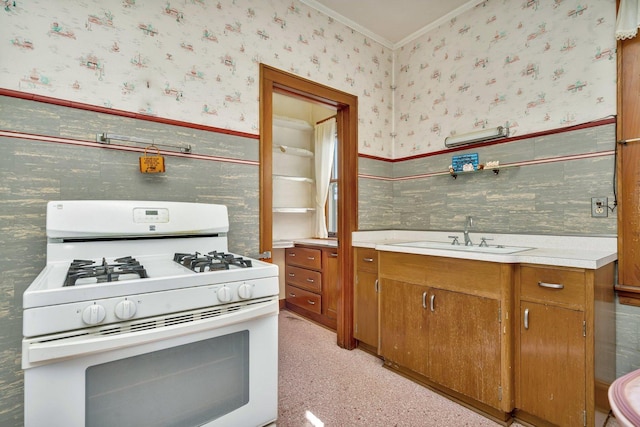 kitchen featuring a wainscoted wall, white gas stove, a sink, brown cabinetry, and wallpapered walls