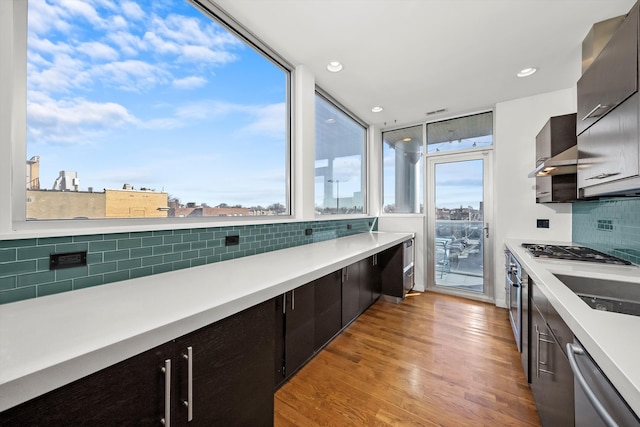 kitchen featuring light wood finished floors, light countertops, appliances with stainless steel finishes, wall chimney exhaust hood, and backsplash