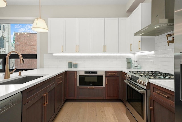 kitchen with a sink, light wood-style floors, light countertops, wall chimney range hood, and appliances with stainless steel finishes