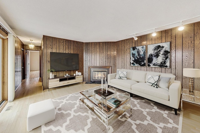 living room featuring rail lighting, visible vents, light wood-style floors, a glass covered fireplace, and wood walls