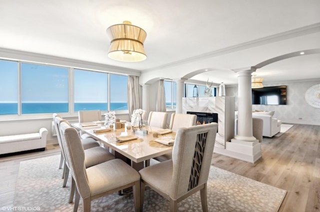 dining room with a water view, light wood-type flooring, crown molding, a fireplace, and ornate columns