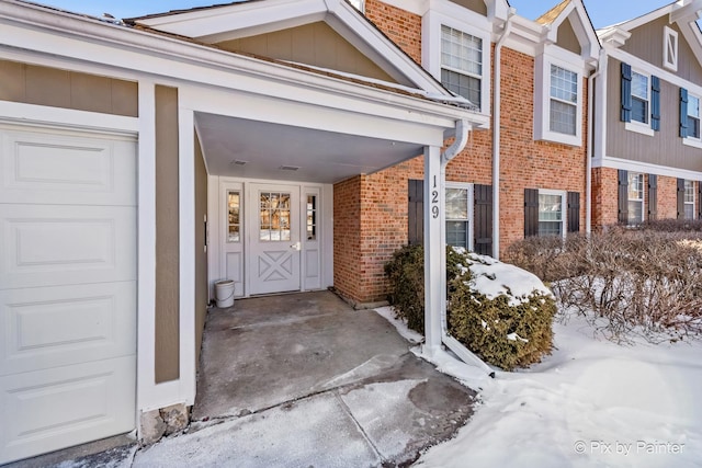 snow covered property entrance with board and batten siding, brick siding, and an attached garage