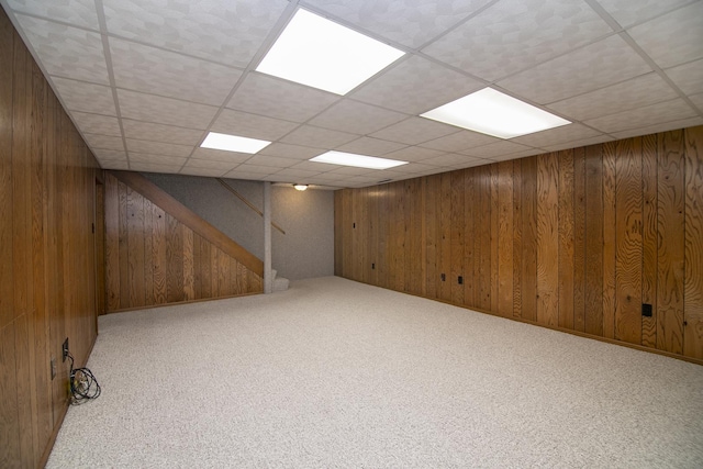 finished below grade area featuring light carpet, stairs, wooden walls, and a drop ceiling