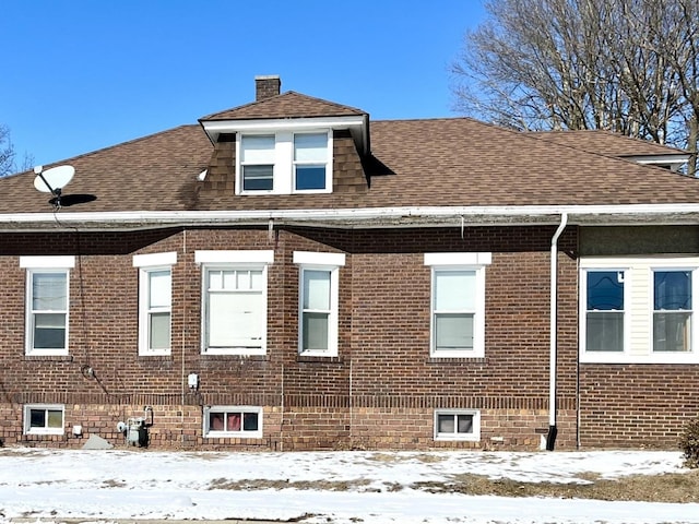 snow covered property featuring a shingled roof, brick siding, and a chimney