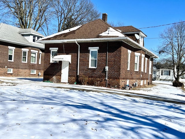 snow covered property with a shingled roof, brick siding, and a chimney