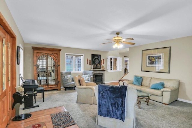 living area featuring a ceiling fan, a glass covered fireplace, carpet, and baseboards