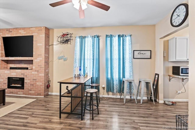 dining area featuring ceiling fan, dark wood-type flooring, a brick fireplace, and baseboards