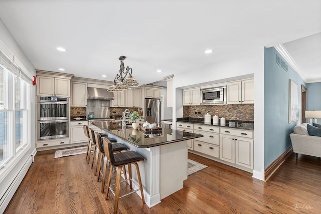 kitchen with pendant lighting, a center island with sink, stainless steel appliances, dark countertops, and a baseboard heating unit