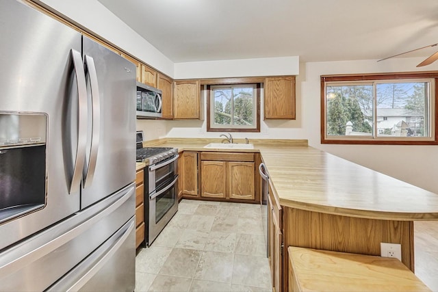 kitchen with a peninsula, a sink, a ceiling fan, appliances with stainless steel finishes, and brown cabinets