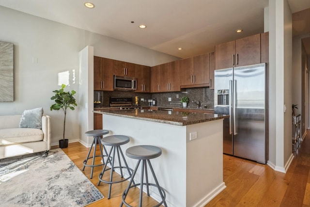 kitchen with stainless steel appliances, backsplash, light wood finished floors, dark stone countertops, and a kitchen bar