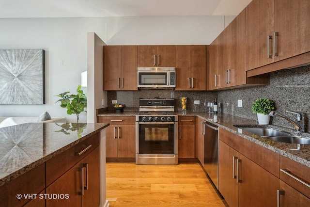 kitchen featuring appliances with stainless steel finishes, light wood-style floors, brown cabinetry, a sink, and dark stone countertops