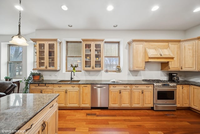 kitchen with pendant lighting, custom exhaust hood, appliances with stainless steel finishes, glass insert cabinets, and a sink