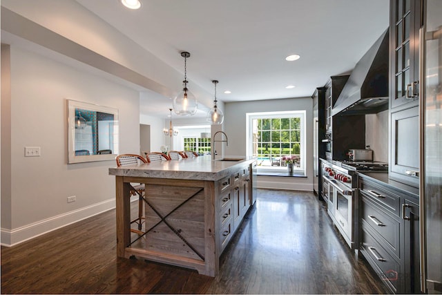 kitchen with a kitchen island with sink, a sink, dark wood-type flooring, appliances with stainless steel finishes, and wall chimney range hood