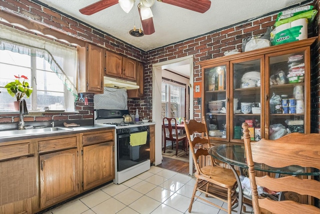 kitchen featuring range with gas stovetop, brick wall, light tile patterned flooring, a sink, and under cabinet range hood