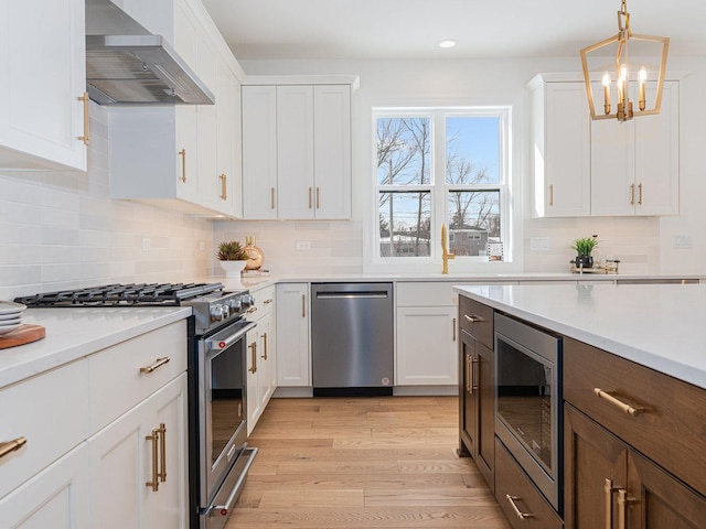 kitchen with wall chimney exhaust hood, stainless steel appliances, white cabinets, light hardwood / wood-style floors, and pendant lighting